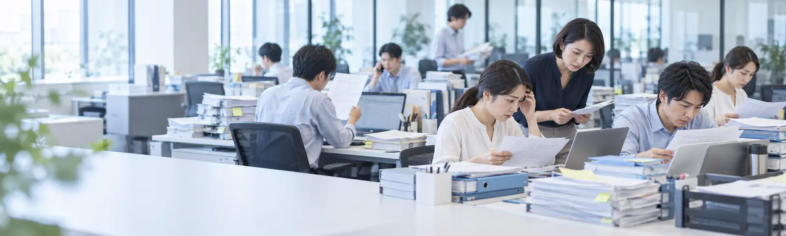 書類やファイルが積まれたオフィスで、社員が忙しく作業する様子